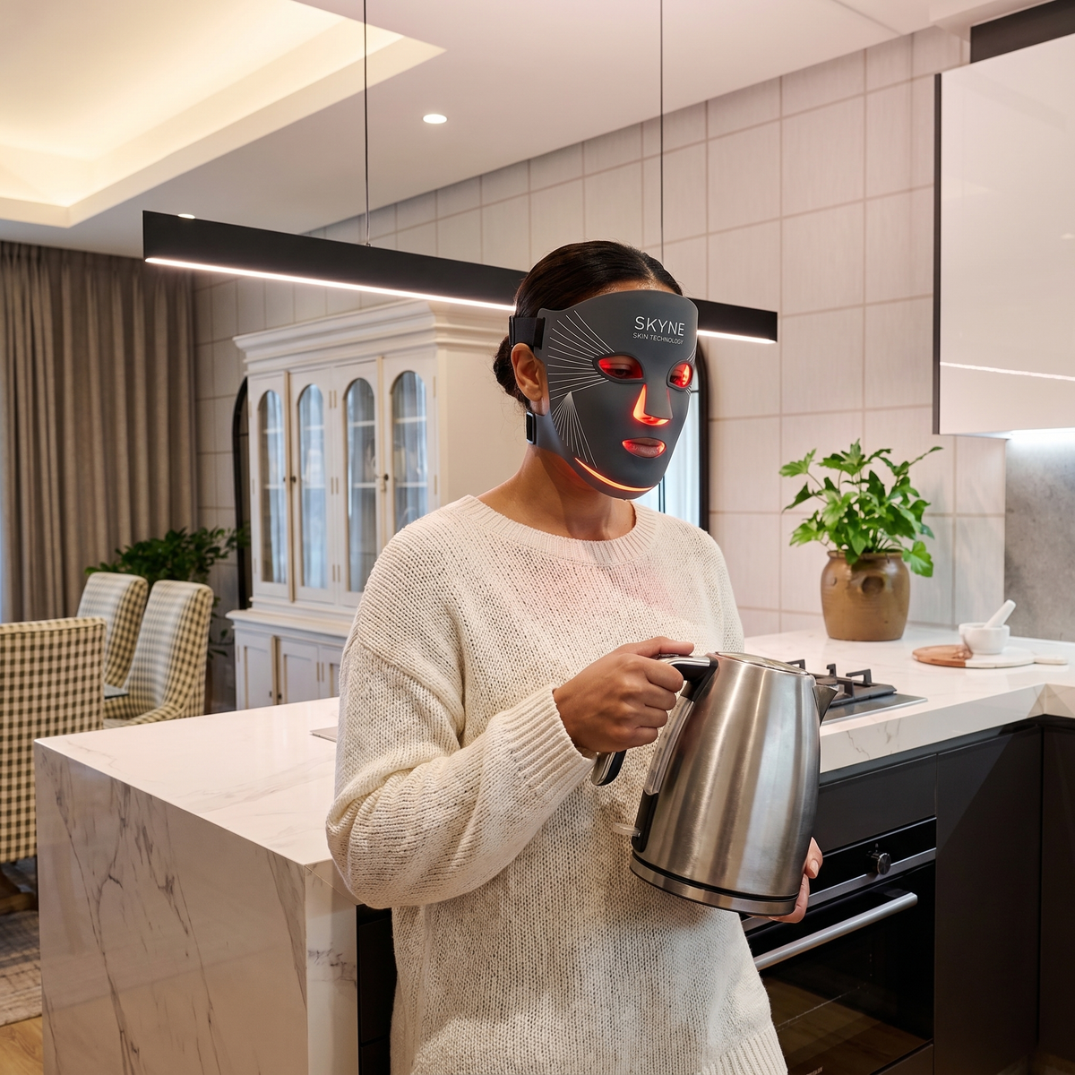 A woman wearing a charcoal LED face mask hands-free while holding a kettle in a kitchen