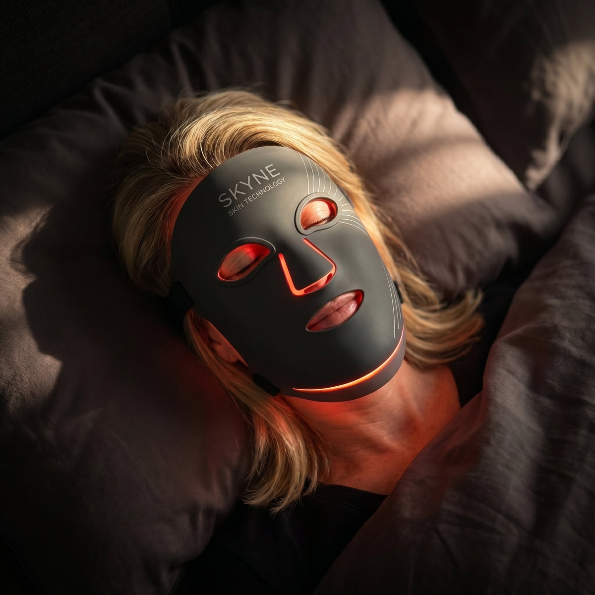 A woman in a charcoal LED mask reclining on a bathroom floor in soft evening light